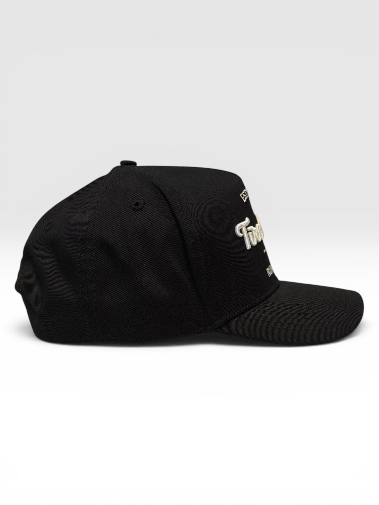 Side view of a black five panel cap with a embroidered logo on the front on a white background